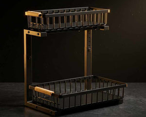 A two-tier wire basket shelf with a black metal body and warm brass-toned frame, featuring light wood handles on the upper basket rim and a lower basket handle, shown against a dark studio background.