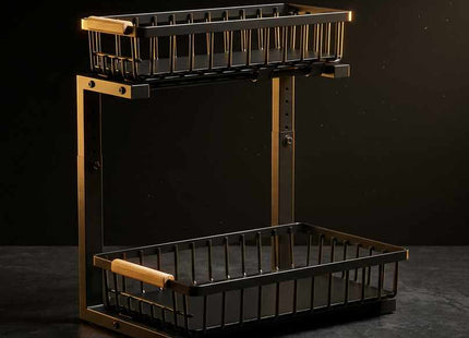 A two-tier wire basket shelf with a black metal body and warm brass-toned frame, featuring light wood handles on the upper basket rim and a lower basket handle, shown against a dark studio background.