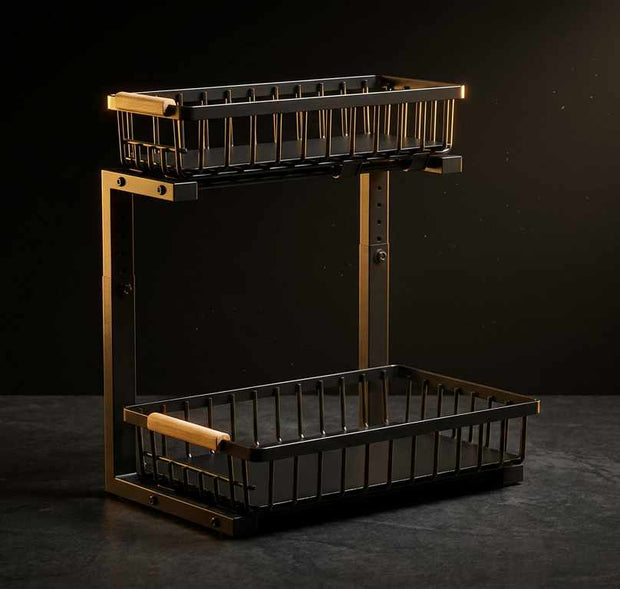 A two-tier wire basket shelf with a black metal body and warm brass-toned frame, featuring light wood handles on the upper basket rim and a lower basket handle, shown against a dark studio background.